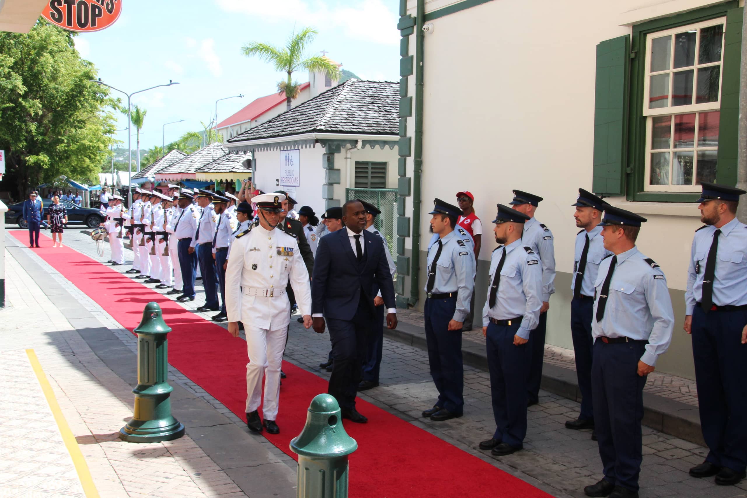 The Governor of Sint Maarten and the aide-de-camp walk along a red carpet, past a guard of honor, to the entrance of the Parliament Building.