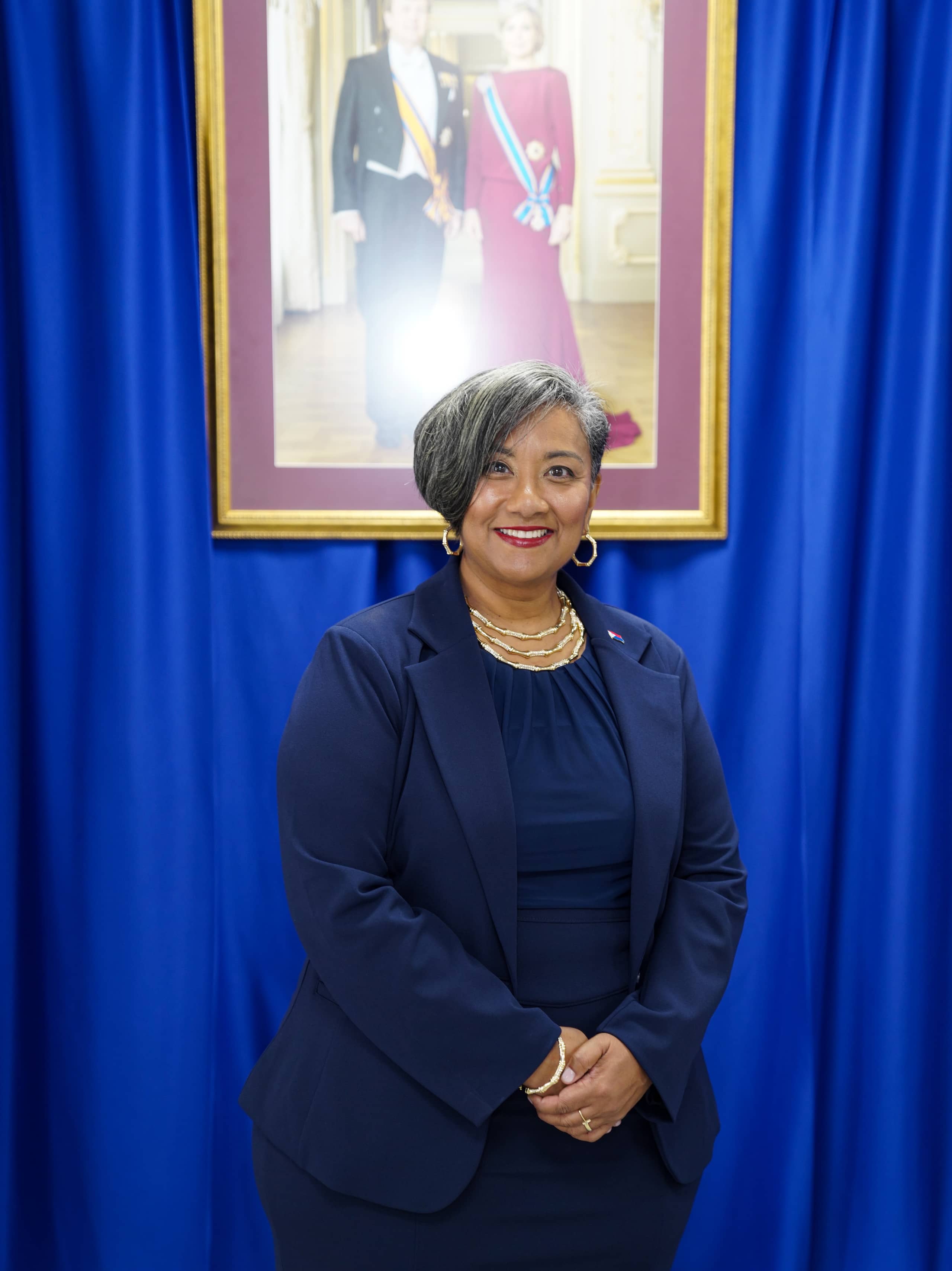 Acting Governor Bird-Lake standing in front of the official portrait of King Willem-Alexander and Queen Maxima.
