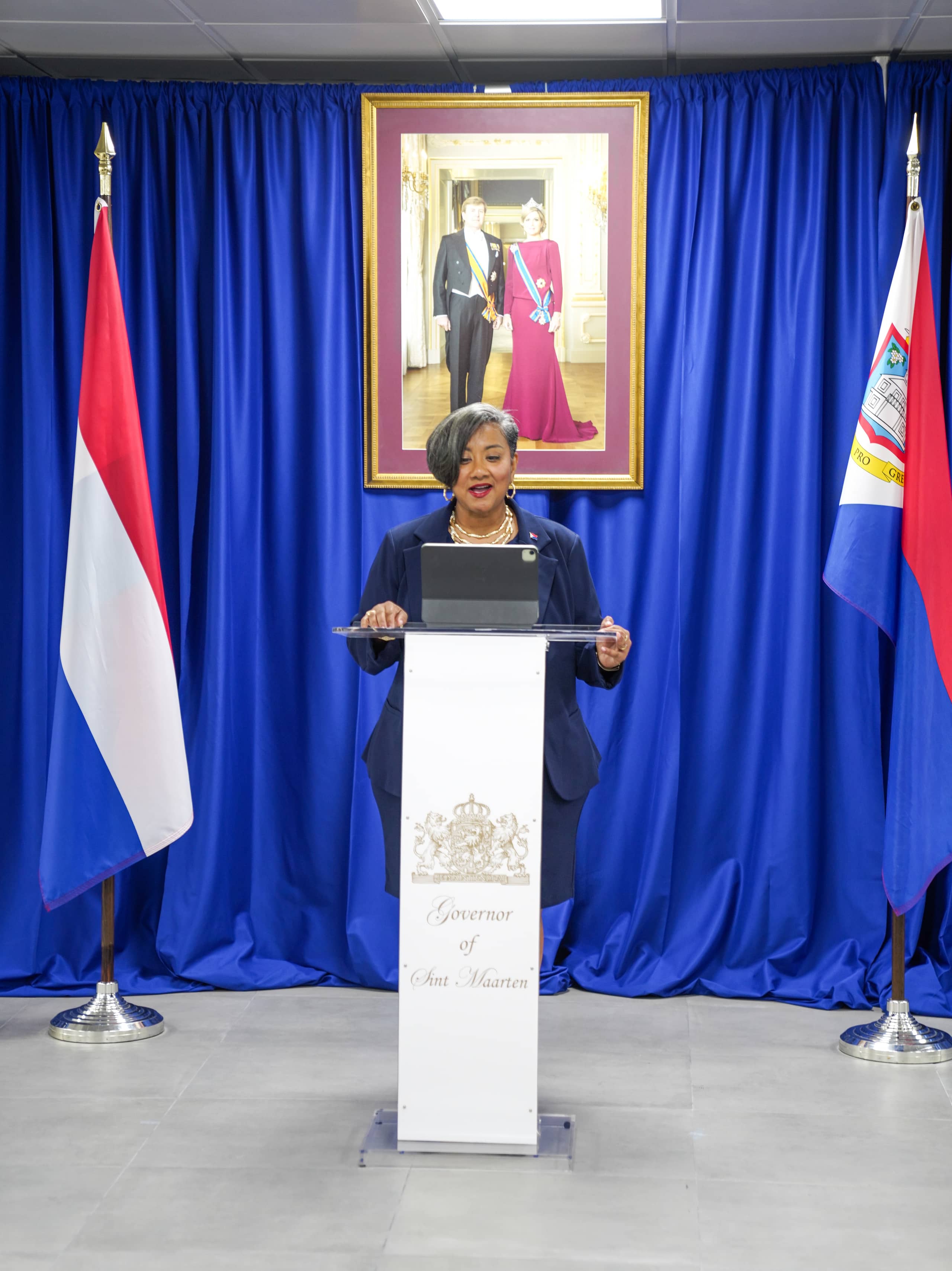 Acting Governor Bird-Lake delivers her speech. In the background is the official portrait of King Willem-Alexander and Queen Maxima. To the left, behind the Acting Governor, stands the Dutch flag; to the right, the Sint Maarten flag.